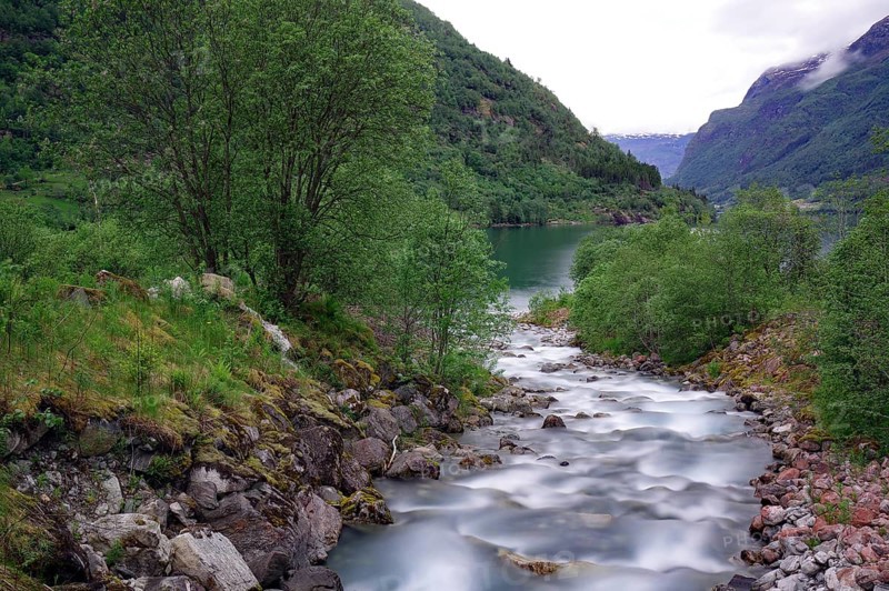 A calm river flows through a green mountain landscape, surrounded by trees and cloudy skies, Olden, Fjordland, norway