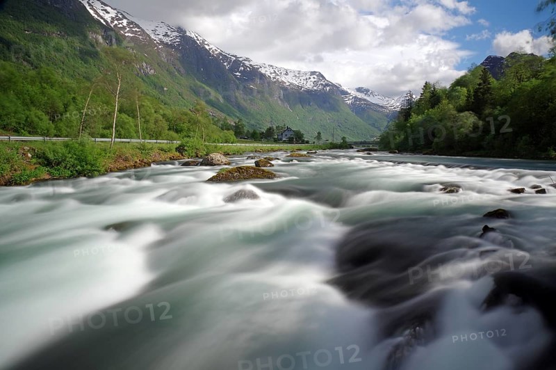A wide river in a mountainous landscape with snow-capped peaks and cloudy skies, Olden, Nordfjord, Norway, Europe