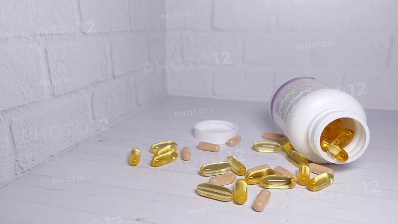 An open bottle of vitamin capsules has spilled yellow and beige pills onto a white table against a white brick wall