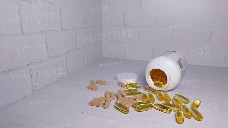 An open bottle with its lid off, spilling out yellow capsules and beige pills onto a light wooden surface against a white brick wall