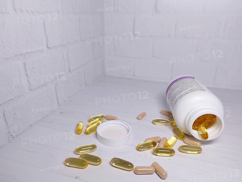 An open supplement bottle on its side with pills and capsules scattered on a white table in front of a white brick wall