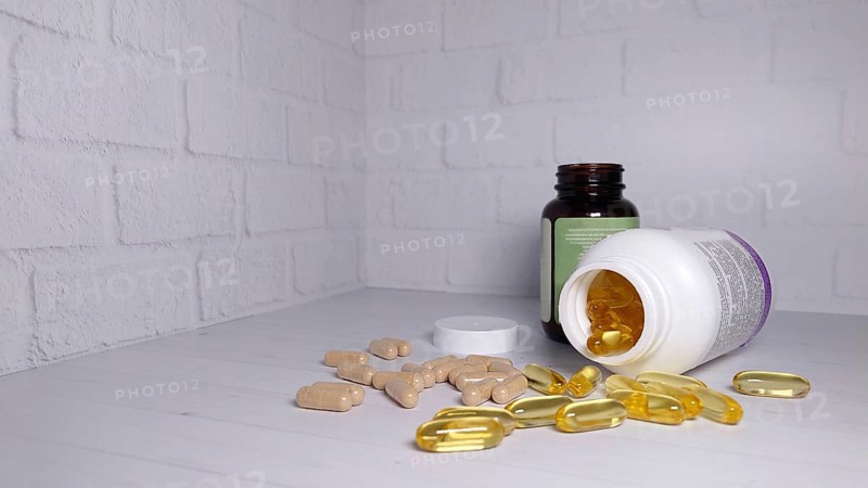 Supplements and pills scattered on a white surface with two bottles, one transparent and one opaque, against a white brick background