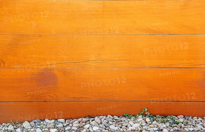 Wooden fence with orange-brown planks, pebbles and rocks at the bottom, a small green plant growing among the stones, background image