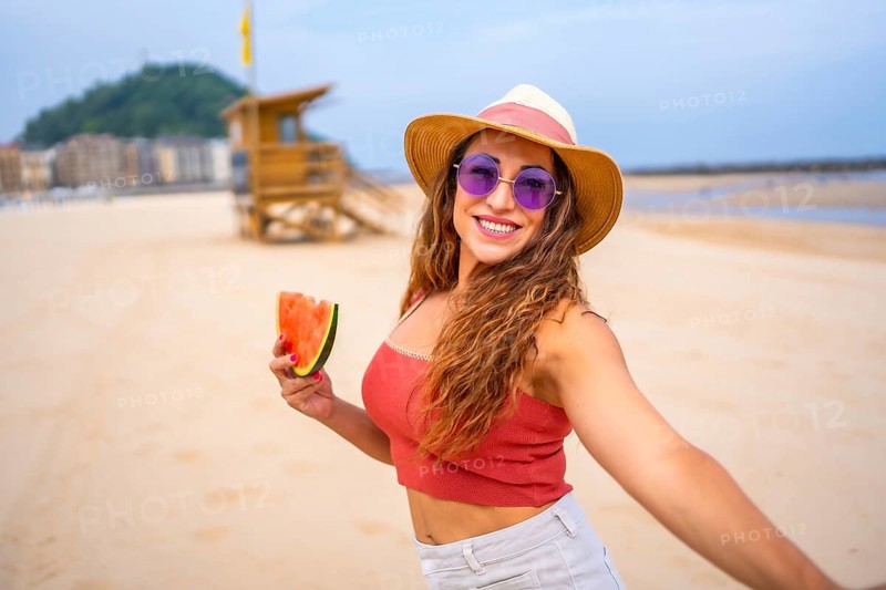 A woman is smiling and holding a watermelon slice. She is wearing a straw hat and sunglasses