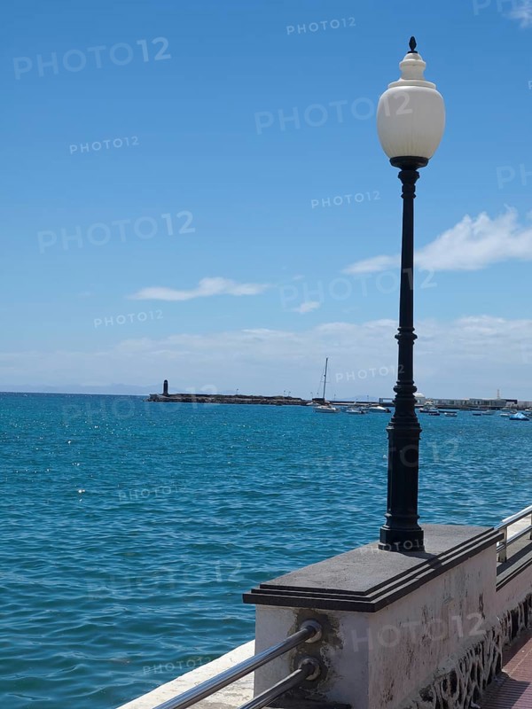 Coastal view of the calm sea with a lantern in the foreground and a harbour in the background under a blue sky, arrecife, Lanzarote, Canary Islands, Spain, Europe