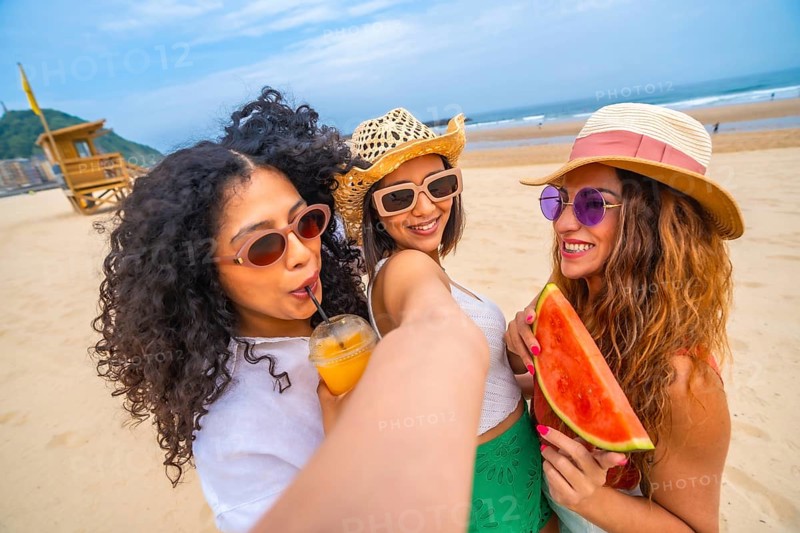 Three women are posing on a beach, one of them holding a watermelon. They are smiling and taking a selfie