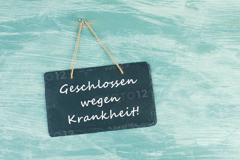 The words closed because of illness are standing in german language on a chalkboard sign, hanging on a blue textured weathered door, concept for business, stores and restaurants