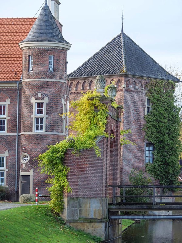 A historic red-brick castle with towers, covered in ivy and surrounded by autumn trees, plus a small wooden bridge, gemen, münsterland, westphalia, germany