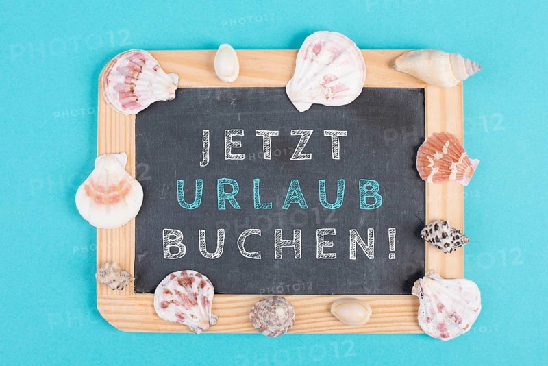 Book vacation now is standing in german language on the chalkboard, seashells bulit frame, holiday and summertime topic, travel concept