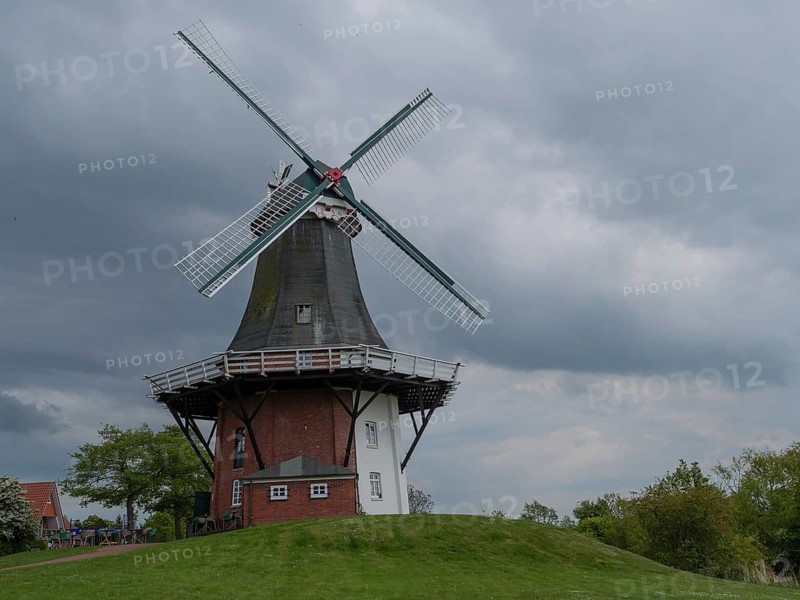 Old windmill on a green hill in front of a cloudy sky and a brick white building, greetsiel, east frisia, germany