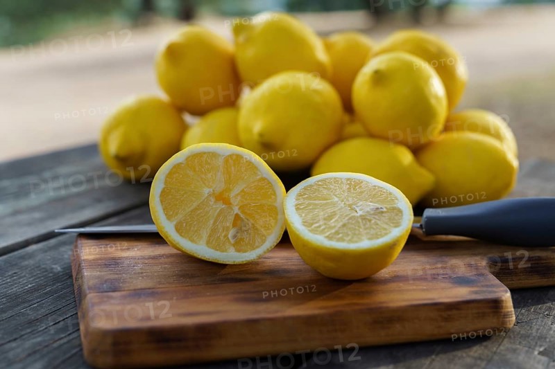 Lemon cut with a knife on a wooden board on a table in a park with a pile of lemons and trees in the background