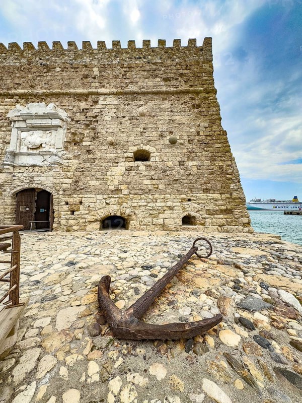 Old historical cane anchor made of iron slightly rusty in front of main gate entrance of fortress built in the Middle Ages 16th century by the Republic of Venice old name Castello Rocca al Mare today Koules in the harbour of Iraklion, right at the edge of the picture harbour basin on Mediterranean Mediterranean coast north coast of island Crete Aegean Aegean Cretan Sea, Heraklion, Crete, Greece, Europe