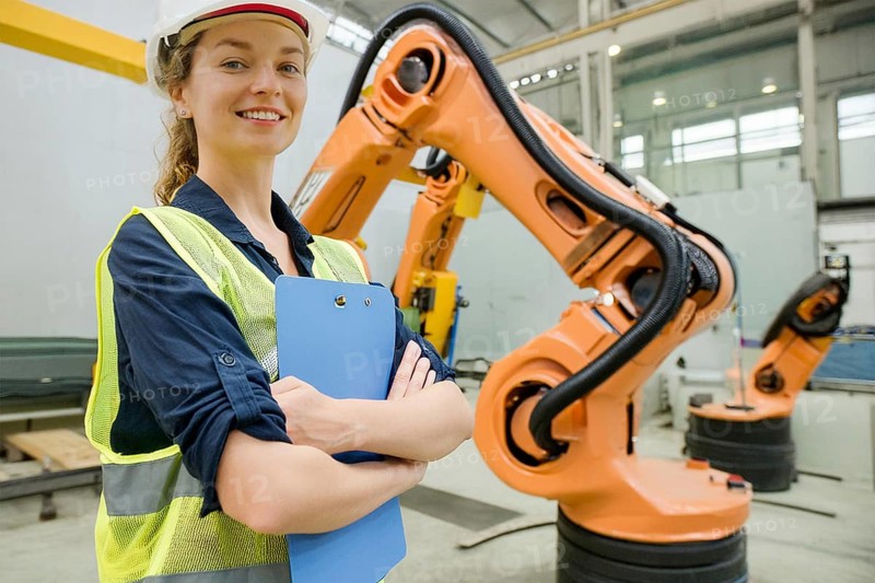 Woman with helmet and yellow safety waistcoat in a factory next to an orange robot arm, AI and illustration technology, AI generated