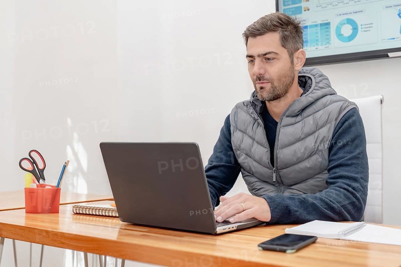An attractive mature man with a shaved beard sits at his desk, deeply focused on his work while looking intently at the screen. The image captures a moment of concentration and professionalism, perfect for business and office themes