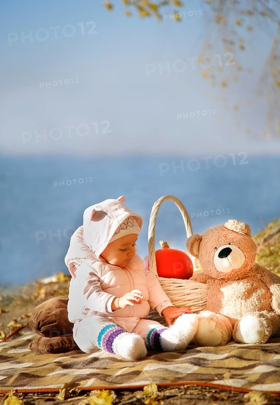 A baby dressed in a pink outfit sits on a blanket near a lake, accompanied by a teddy bear and a basket with a pumpkin.Belarus.Minsk