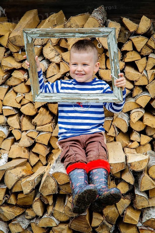 A smiling boy in a striped shirt and boots sits on a woodpile, holding a rustic, weathered frame around his head.Belarus.Minsk