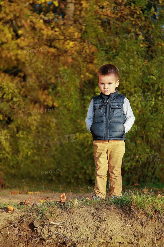 A young boy in a blue vest stands on a grassy area surrounded by autumn leaves and trees, Belarus.Minsk