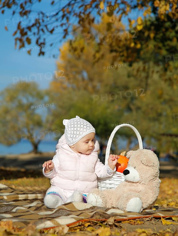 Baby in a pink jacket and white hat sitting on a blanket with a teddy bear and basket in an autumn park, Belarus.Minsk