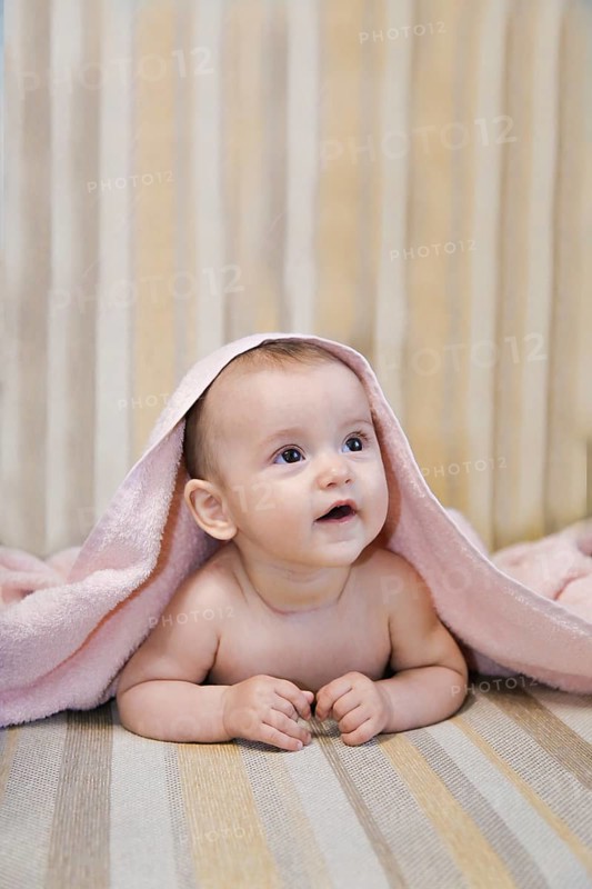 A baby lying on a striped surface, partially covered with a pink towel, looking up with an adorable expression