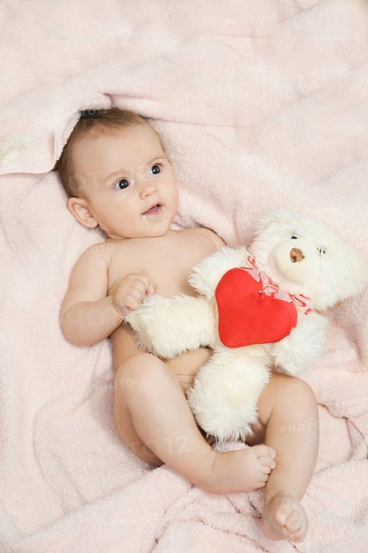 A baby lying on a pink blanket holding a white teddy bear with a red heart