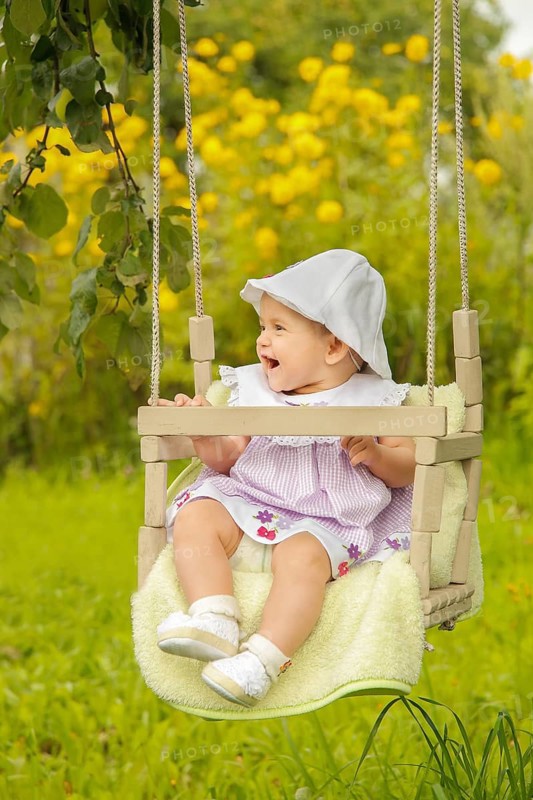 A baby sitting on a swing in a lush garden with yellow flowers, wearing a hat and dress while smiling, Belarus.Minsk
