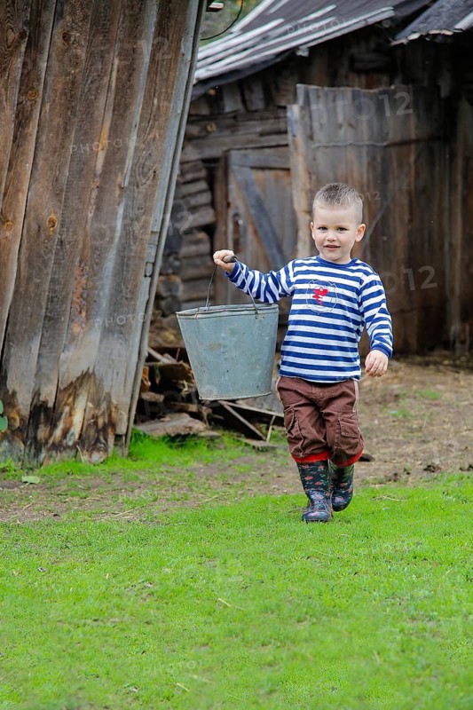 A boy carrying a metal bucket, wearing a striped shirt, brown pants, and rubber boots, walking on grass in a rustic, rural setting.Belarus.Minsk