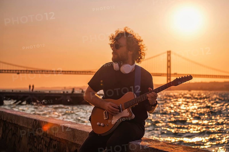 Hipster street rock musician in black playing electric guitar in the street on sunset on embankment with 25th of April bridge in background. Lisbon, Portugal, Europe
