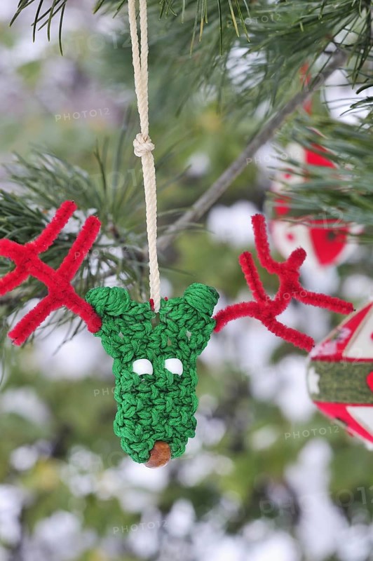 Funny green and red handmade reindeer Christmas ornament with pipe cleaner antlers and other red and white felt ornaments hang from the boughs of an Austrian pine tree (pinus nigra) outdoors with snow on the branches and copy space for a greeting card or other text. Vertical. Selective focus