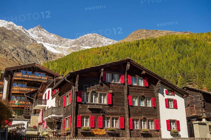 A traditional house with red shutters against a backdrop of forest and mountains in autumn, Saas Fee, Switzerland, Europe