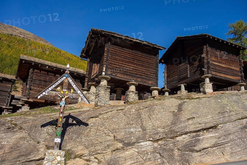 Traditional wooden houses and a cross on a rocky mountainside under a blue sky, Saas Fee, Switzerland, Europe