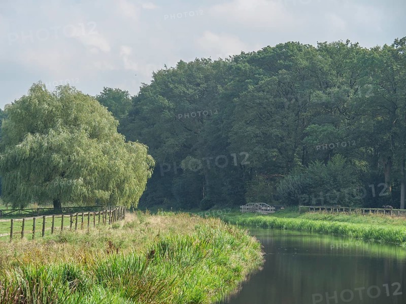 A calm river flows along a wooded bank and a green meadow under a cloudy sky, haaksbergen, . netherlands