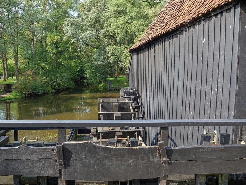 A historic wooden building next to a river and a bridge, surrounded by dense forest, haaksbergen, . netherlands