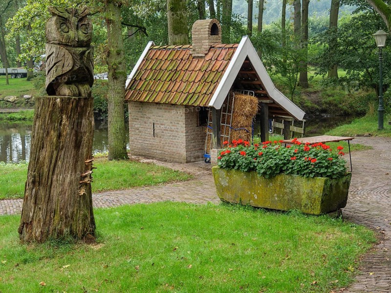 A rural garden with a carved wooden owl, a small brick house, a profusion of flowers and old trees, haaksbergen, . netherlands
