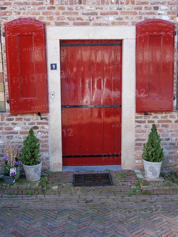 Red door with shutters surrounded by brick wall and potted plants, Doesburg, Netherlands