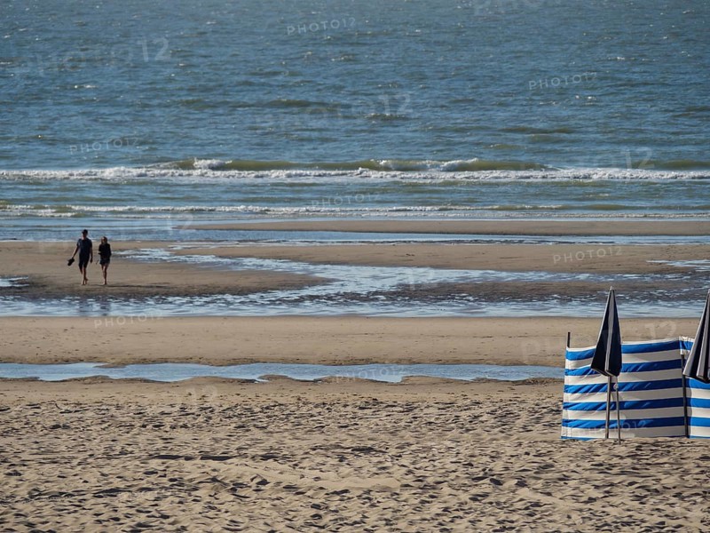 A beach with sand and sea, two people walking along the shore. Blue and white parasols and a quiet atmosphere, DE Haan, Belgium, Europe