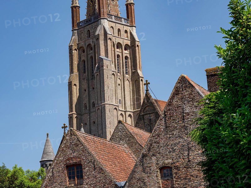 A tall brick church tower next to several historic buildings with Gothic architecture under a blue sky, Bruges, Belgium, Europe