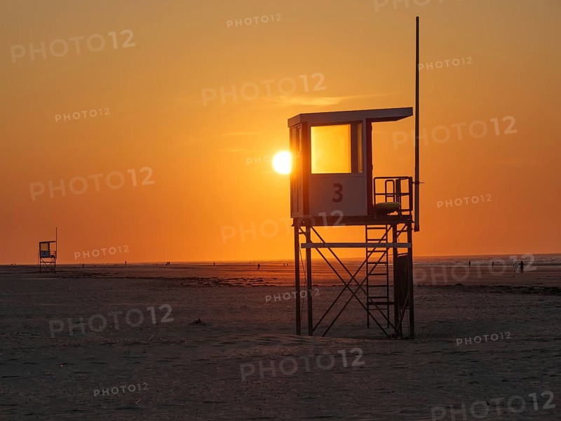 A rescue tower stands on the beach in front of a bright orange sunset, the sea in the background, juist, east frisia, germany