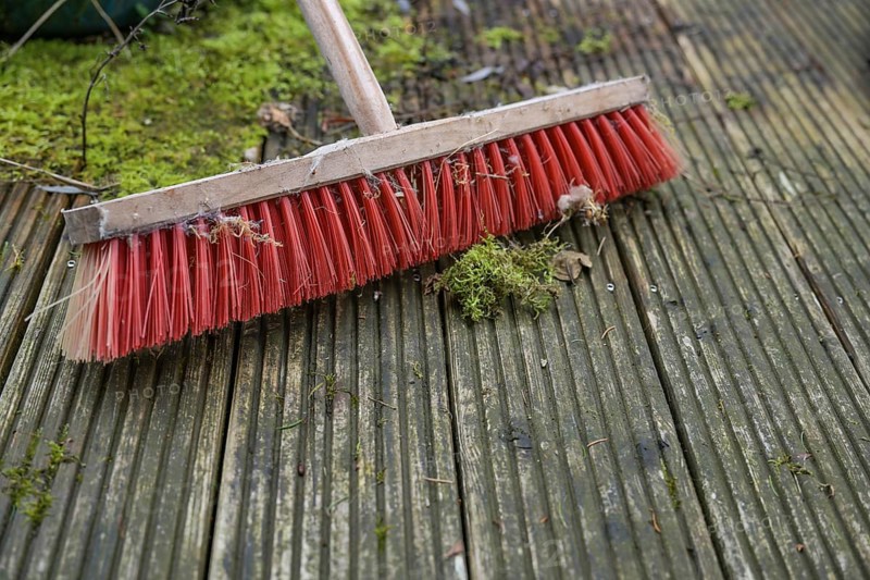 Outdoor broom with red plastic bristles on a weathered wooden deck with slippery algae and moss, spring cleaning in garden and yard, copy space, selected focus, narrow depth of field
