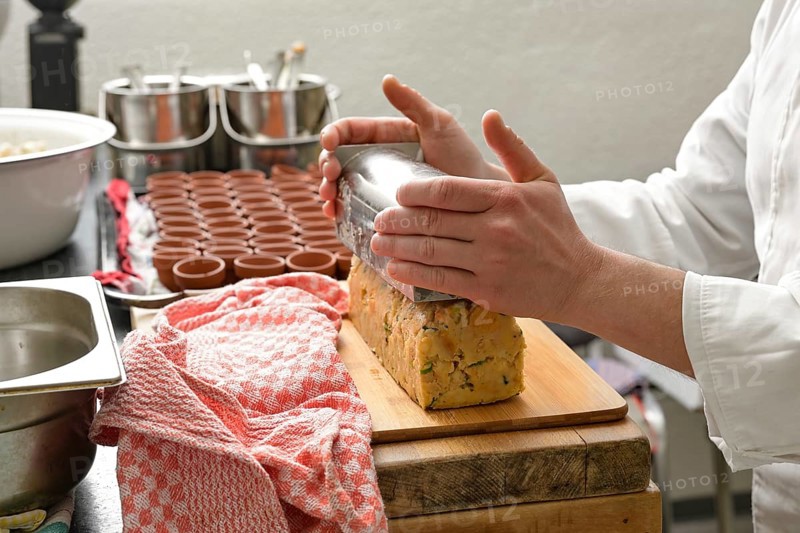 Hands of a cook toppling a savory bread pudding from a stainless steel mold onto a wooden board in a professional kitchen, selected focus, narrow depth of field