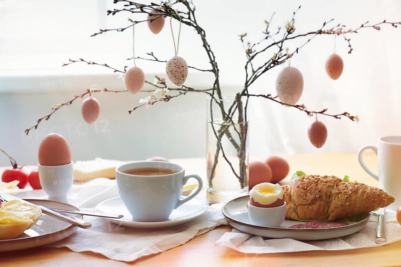 Easter breakfast in the morning sun with coffee and croissant on a table at the window, decoration from natural eggs hanging on spring branches, copy space, selected soft focus, narrow depth of field