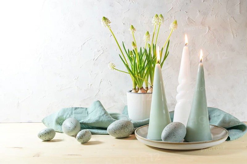 Easter eggs and candles in pastel turquoise color and a potted white grape hyacinth on a wooden table against a light plastered wall, holiday decoration, greeting card, copy space, selected focus, narrow depth of field