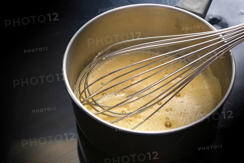 Preparation of hollandaise sauce, egg mixture is whisked at low temperature until foamy in a steel pot on a black cooktop, cooking concept, copy space, selected focus, narrow depth of field