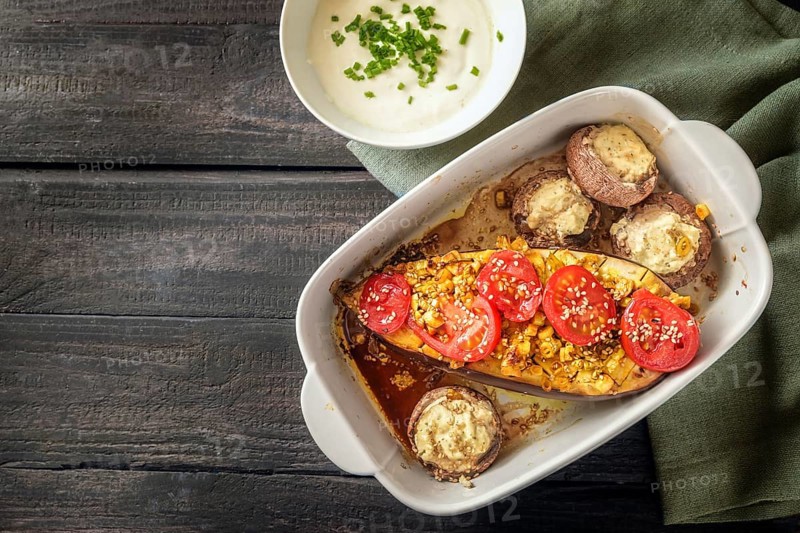 Baked eggplant and mushrooms with garlic, ginger, onions, tomatoes and sesame seed in a casserole, served with yoghurt on a napkin and a dark rustic wooden table, copy space, top view from above