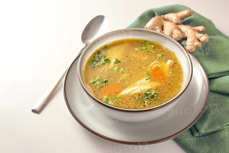 Homemade chicken soup with vegetables, ginger and parsley in a bowl, green napkin and spoon on a gay white table, copy space, selected focus, narrow depth of field