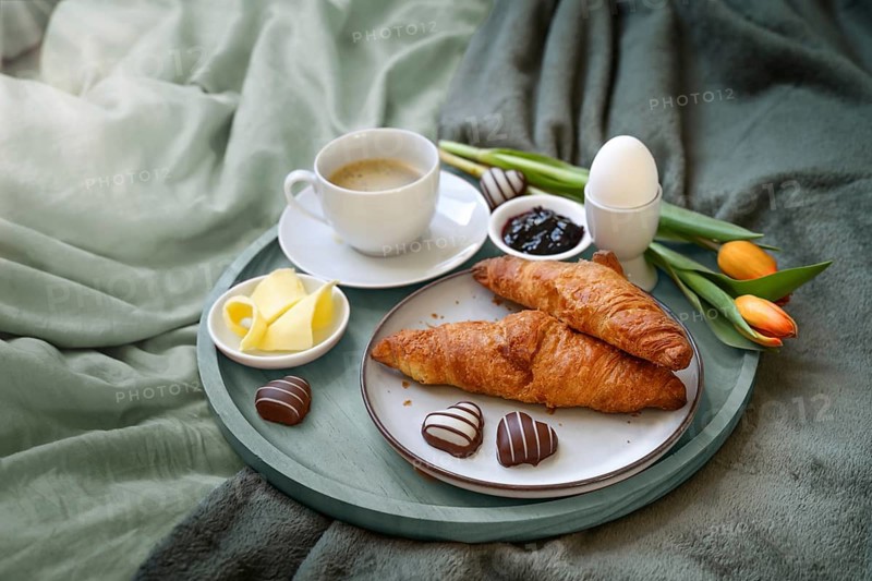 Breakfast served with love in bed, coffee, croissant, jam, egg, some chocolate hearts and tulips on a tray on gray green bed linen at a romantic holiday morning, copy space, selected focus
