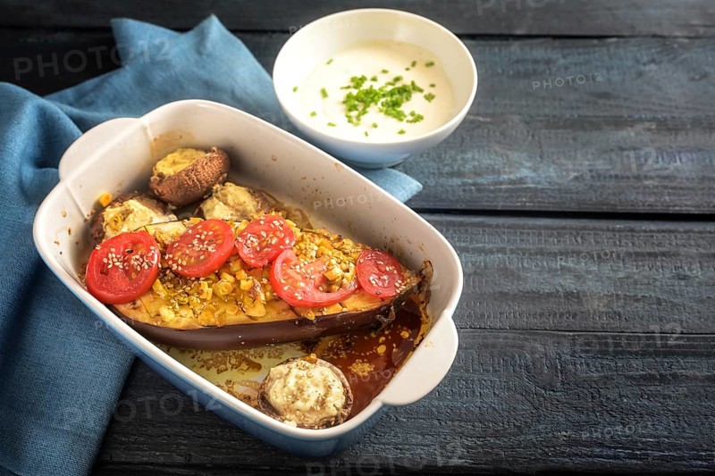 Baked half eggplant and stuffed mushrooms with garlic, ginger, onions and tomatoes in a casserole, served with yoghurt dip on a blue napkin and a dark rustic wooden table, copy space, selected focus