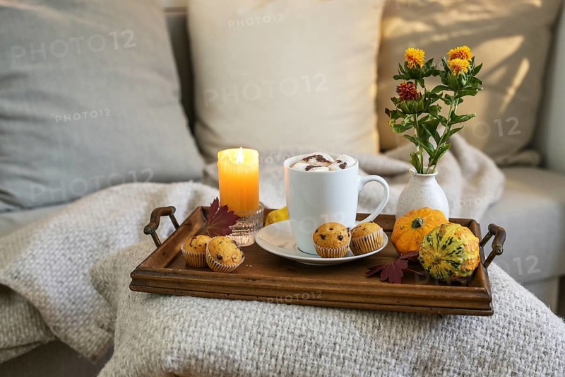 Hot chocolate drink with marshmallows and mini muffins on a tray with candle, flowers and autumn decorations beside a cozy couch, copy space, selected focus, narrow depth of field