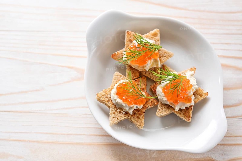 Canapes with red caviar on toasted bread in star shape with cream and dill garnish on a white plate, light wooden table, copy space, high angle view from above, selected focus