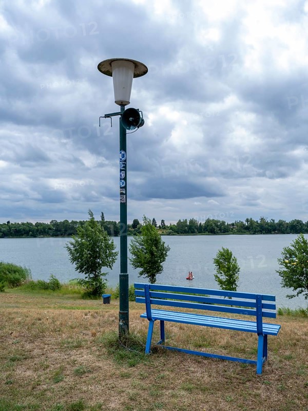 Weathered street lamp with loudspeakers next to a blue wooden bench at Lake Jersleben, Jersleben, Saxony-Anhalt, Germany, Europe