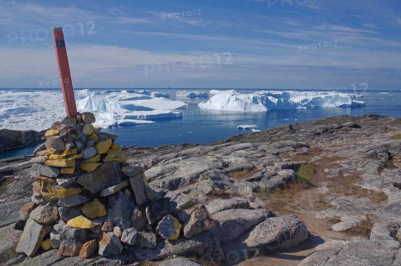 A pile of stones with a flag on rocky terrain in front of an arctic landscape and icebergs in the water, signpost, Ilulissat, Disko Bay, Greenland, Denmark, North America